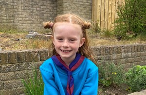 Photo shows young girl with red hair in buns wearing a blue jumper, red and blue scout tie and purple and black spotted trousers