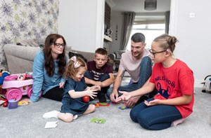 A family of five sit on the carpet playing a game together.