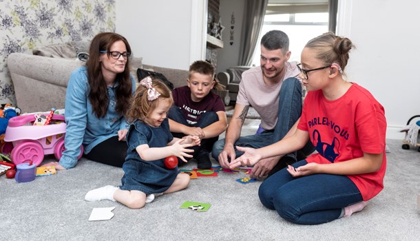 A family of five sit on the carpet playing a game together.