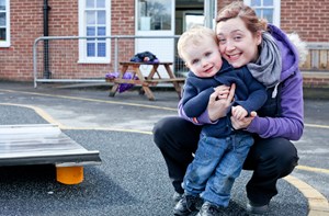 A woman crouches on the floor, holding a toddler outside a care setting.