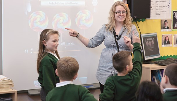 A teacher in a primary school has a listening loop around her neck and points to the projector screen. 