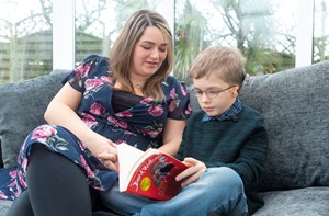 A child reads a book with his mum's help while sitting on the sofa. 