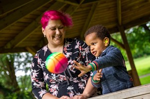 A mum with pink hair smiles by her young son wearing hearing aids and throwing a ball.