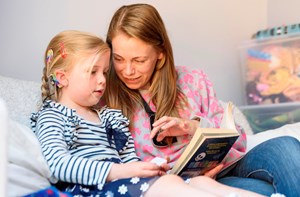 A mum wearing a radio aid reading a book with her young daughter with rainbow cochlear implants on the sofa. 