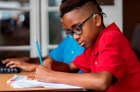 A young black boy with a cochlear implant and glasses studying at a desk