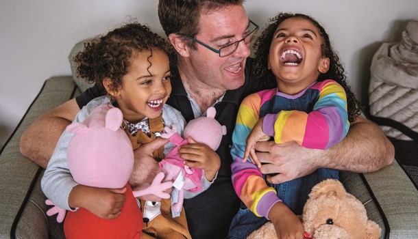 A dad sat on a chair tickling his two young daughters who are holding teddies.