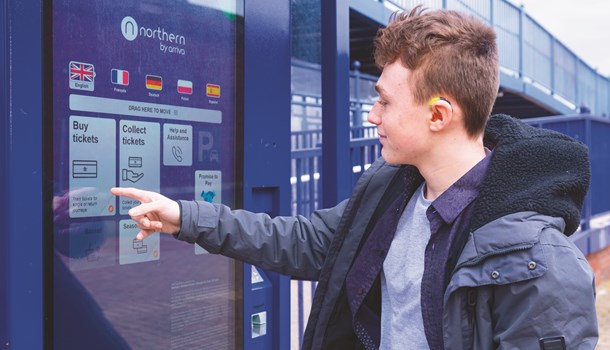 A young man wearing a hearing aid points to a travel sign at a rail station.