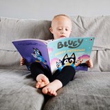 A deaf boy with Down's Syndrome wearing hearing aids and sat on a sofa reading a Bluey children's book.