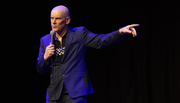 Steve Day doing a stand-up performance with a mic and pointing with his left arm.