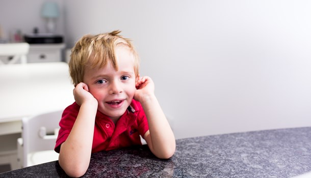 A boy in a red t-shirt leans on a counter with his elbows and has his hands over his ears.