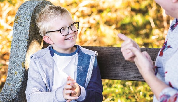 Xander wearing cochlear implants and glasses signing with his mum on a park bench.