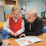 Anna wearing a red school uniform reading a book and practising BSL signs with her dad.