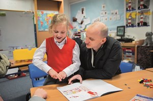 Anna wearing a red school uniform reading a book and practising BSL signs with her dad.