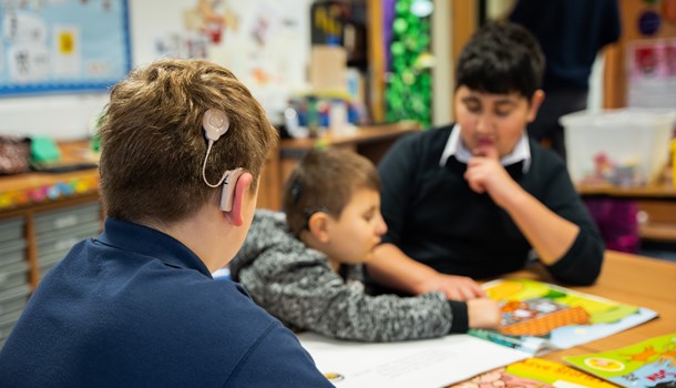Back of a boy's head showing his cochlear implant while sat across from two boys at a classroom table.