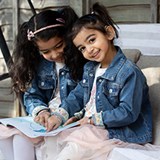 Two young children wearing denim jackets sitting on a lounger in the garden and pointing to a book. One child is facing the camera and smiling