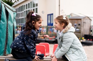 Two girls sit on a bench in a playground, smiling at each other. 