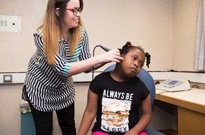 An audiologist stood using an implement in a sitting girl's ear.
