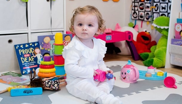 A baby girl in a white romper sat playing on the floor and looking at the camera.