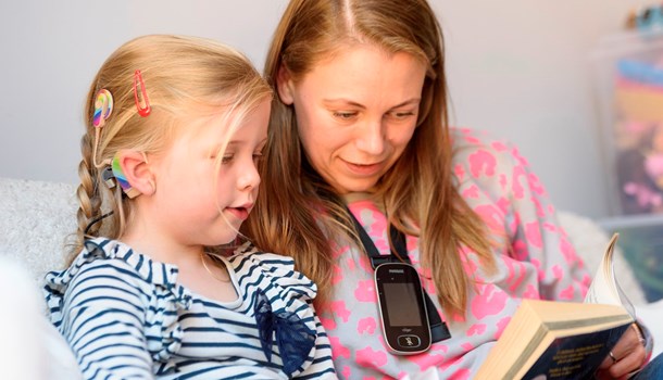 A young deaf girl with a rainbow cochlear implant reading with her mum who's wearing a radio aid.