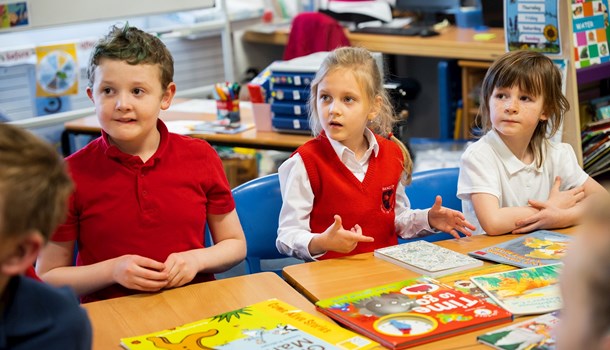 A boy and two girls in uniform sat at a school desk with the middle child signing to the teacher.