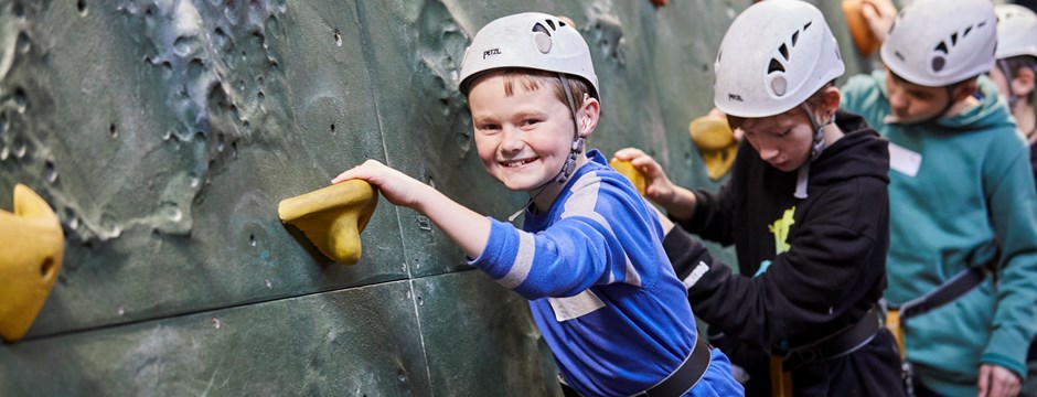 A boy wearing a blue top and a helmet is climbing along a climbing wall. He is smiling at the camera.