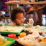 Toddler wearing pink glasses playing with white-coloured foam in colourful trays. Some of the foam is on the child's face and hands