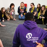 A group of young people sat on the floor in a circle. The person in the middle of the photo has their back to the viewer and is wearing a purple Deaf Children's Society hoodie with the charity's logo on the back