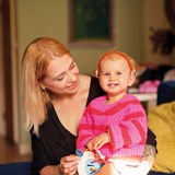 Goldie, aged one, is wearing a bright pink striped jumper, an orange hairband with black dots and hearing aids. She's holding a toy while sitting on her mum's lap