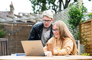 Two young people sat outside in a garden by a table and looking a laptop computer screen