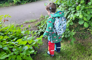 A little boy wearing a tractor-themed raincoat and backpack faces away from the camera. He's wearing cochlear implants.